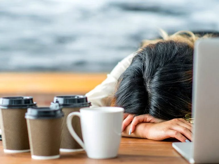 Woman exhausted, with head on desk, surrounded by coffee cups